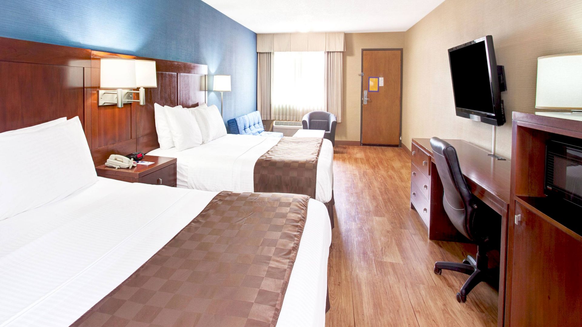 A hotel room with two beds, a flat-screen TV, a desk with a chair, a microwave, and a door leading to an entrance or bathroom.