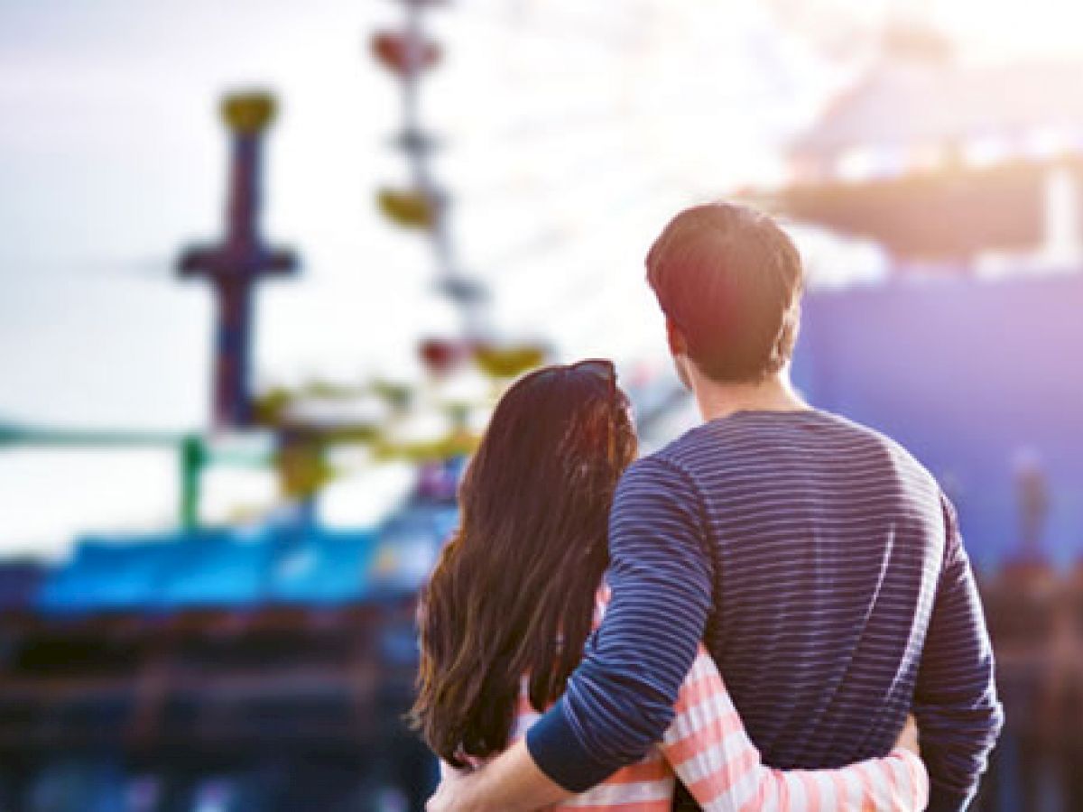 A couple stands with their arms around each other, gazing at a fairground with a Ferris wheel and other amusement rides in the background.