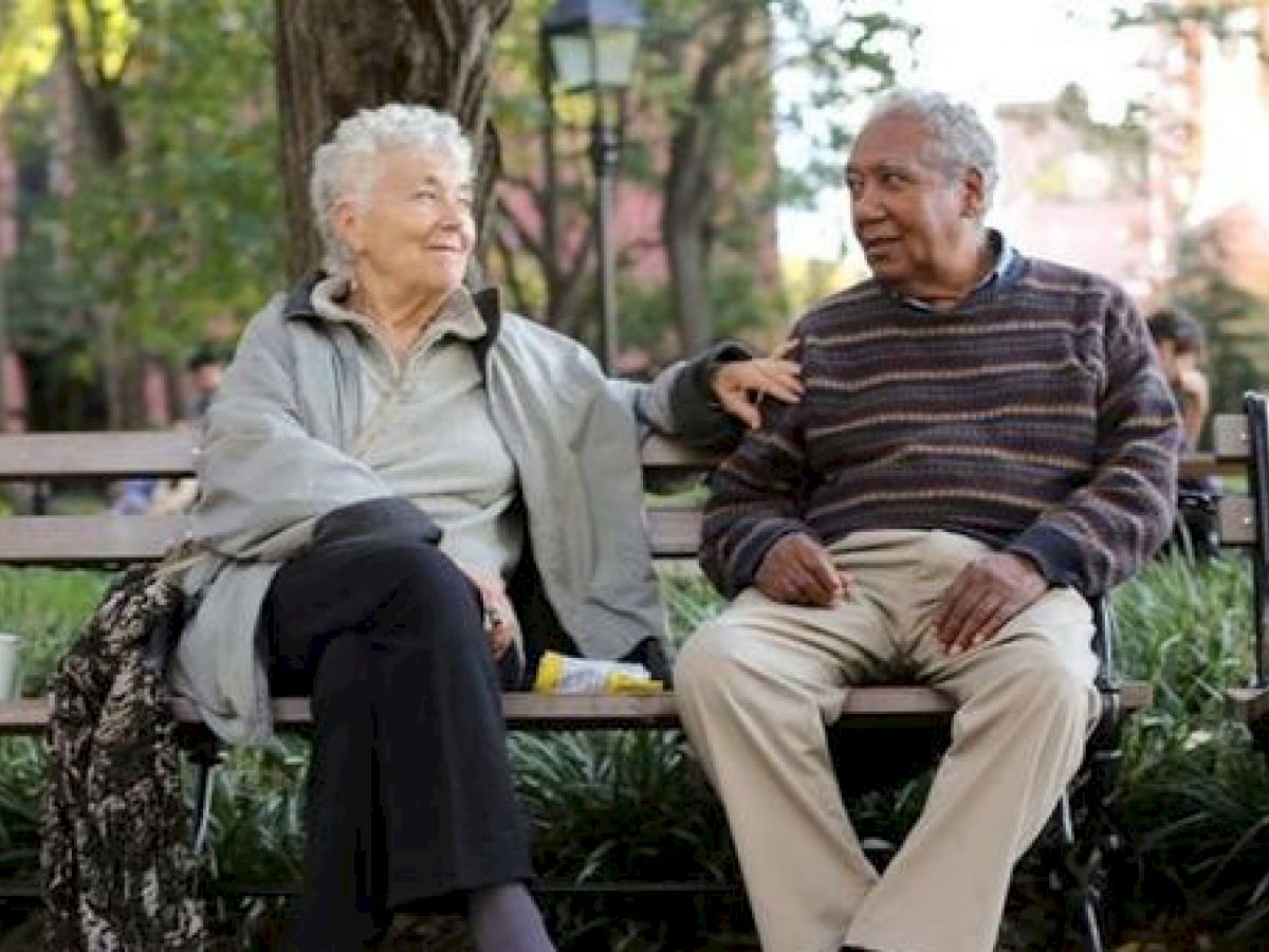 An elderly couple sitting on a bench in a park while having a conversation. There are two cups and a walker placed nearby, with trees in the background.