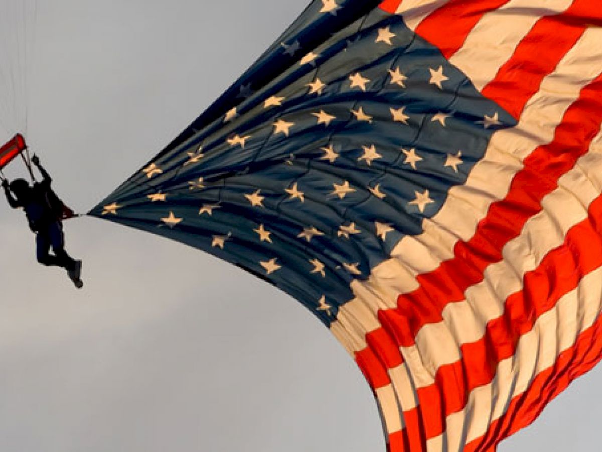 A person is parachuting while holding a large American flag, which is unfurled and prominently displayed in the sky.