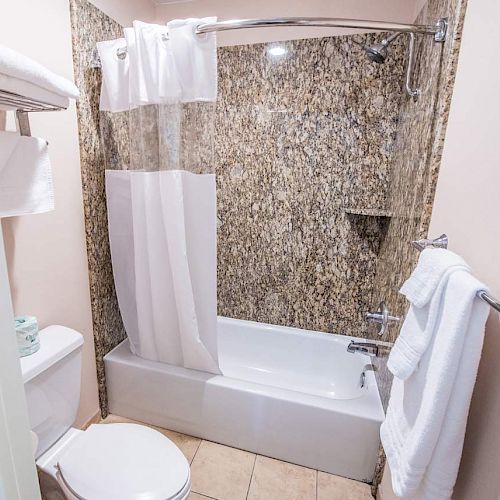 A bathroom with a tub-shower combo, patterned tile walls, white towels, a white toilet, and a towel hanging on a towel bar ends the sentence.