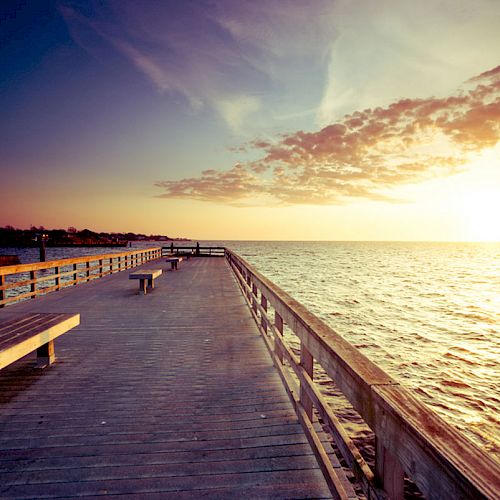 A wooden boardwalk extends over the ocean, bathed in the warm hues of a sunset, with benches along the path and clouds scattered in the sky.