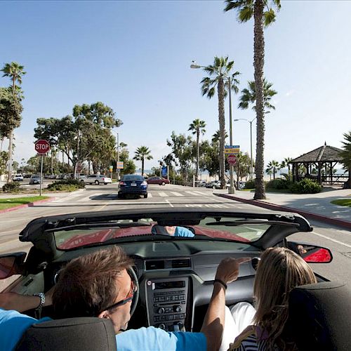 A man and woman drive a convertible along a scenic, palm tree-lined street near the beach on a sunny day, with other cars and people around.