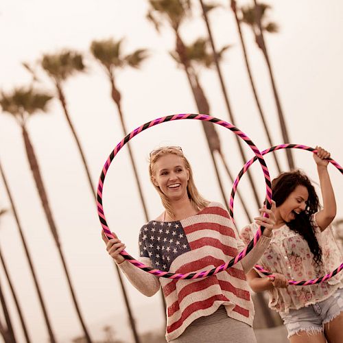 Two women are standing outdoors with palm trees in the background, holding and playing with hula hoops while smiling.