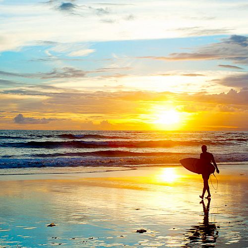 A person is carrying a surfboard along a beach at sunset, with the sky and ocean creating a serene and colorful backdrop.