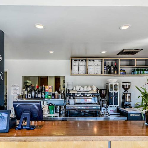 A coffee shop counter with a cash register, coffee machines, menu on chalkboard, tech devices, and plant decor, shelves stocked with items in the background.