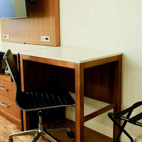 The image shows a hotel room corner with a desk, a black office chair, a TV mounted on the wall, a cabinet below the TV, and a luggage rack.