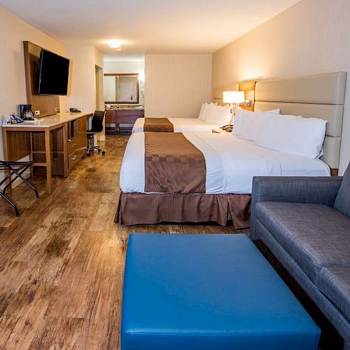 A spacious hotel room with two beds, a TV, a desk area, a sofa, a blue ottoman, and wooden flooring to give it a cozy and modern feel.