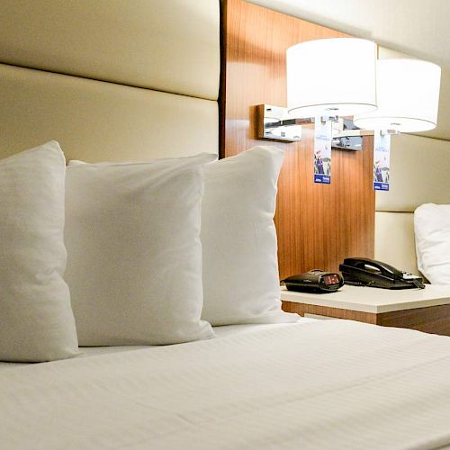 A neatly made bed with white pillows and a headboard, accompanied by a bedside table holding a phone, alarm clock, and two wall-mounted lamps.