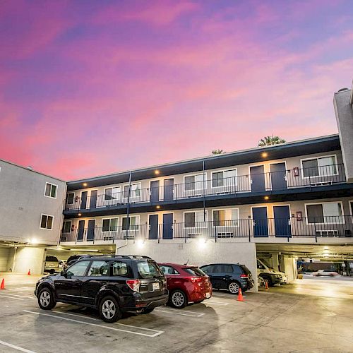 The image shows an apartment complex with multiple floors and a parking area below; cars are parked, and the sky is a colorful purple and pink.