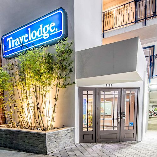 This image shows the entrance of a Travelodge hotel, featuring a neon sign, plants, and glass doors.