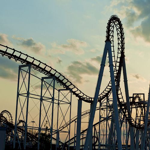 The image shows a large, intricate roller coaster structure against a sunset sky, featuring multiple loops and steep tracks, creating a thrilling scene.