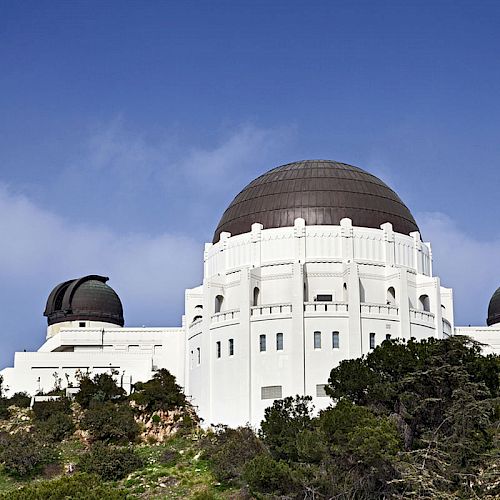 The image shows a white observatory with three domes, situated on a hill among greenery, under a clear blue sky.