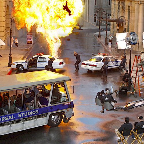 A movie set scene with an explosion, police cars, crew members, and a Universal Studios tour tram filled with people observing the action.