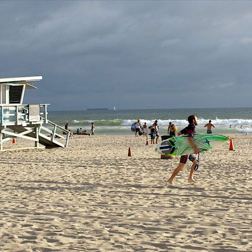 A beach scene with a lifeguard tower, a surfer carrying a board, and several people by the shoreline with cloudy skies in the background.