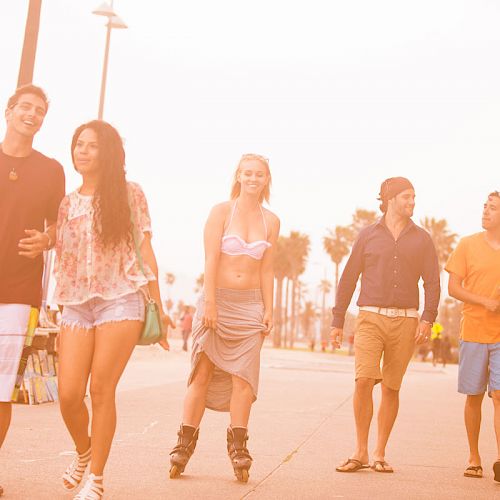 A group of five people, three men and two women, walk and roller skate along a palm-lined beach boardwalk under a warm, golden sunset.