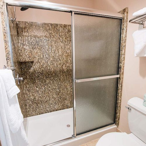 A bathroom with a sliding shower door, white towels on racks, granite shower walls, and a white toilet with a shelf above.