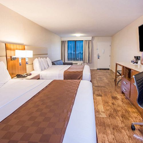 A hotel room with two beds, a work desk, a chair, a mounted TV, and a window. The floor is wooden, and there are lamps by the bed.