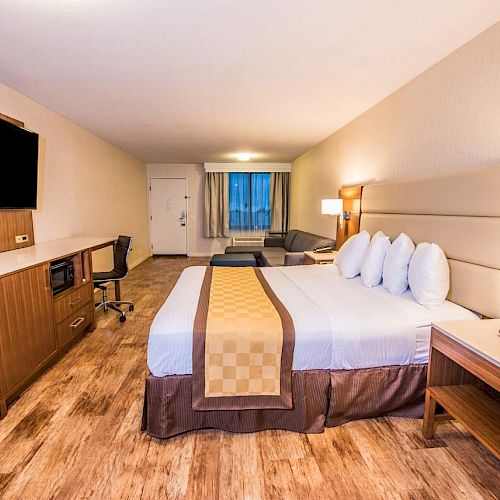 A spacious hotel room with a large bed, a desk, a flat-screen TV, a sofa, and wooden furniture, creating a warm and inviting atmosphere.