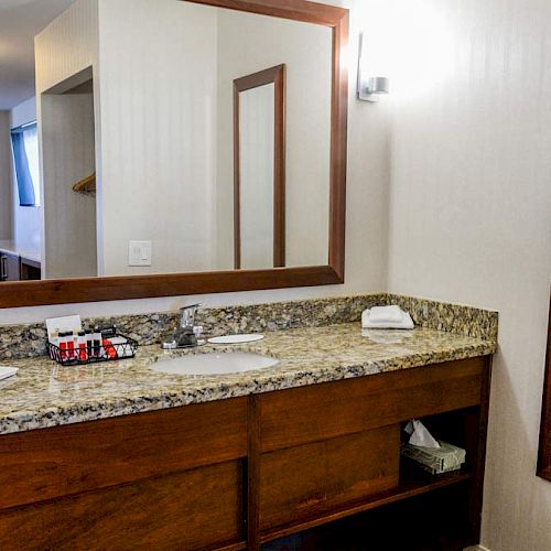 A hotel bathroom with a granite countertop, two mirrors, a hairdryer, tissue box, and toiletries near the sink, with an adjoining room visible.