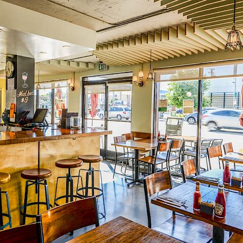 The image shows the interior of a modern, well-lit café with wooden tables, bar stools, and large windows overlooking the street, creating a cozy atmosphere.