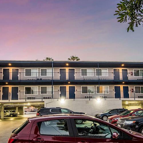 The image shows an apartment building with outdoor entrances, a parking lot with cars, and a purple and pink sky at dusk.