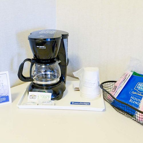 The image shows a coffee station with a coffee maker, coffee packets, disposable cups, and a small sign, all arranged neatly.