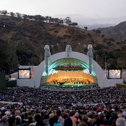 The image shows a large outdoor amphitheater with an audience watching a performance on stage, set against a backdrop of hilly terrain.