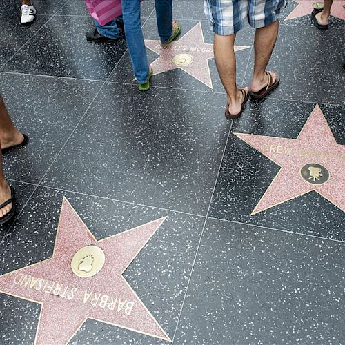 The image shows people walking on a sidewalk with stars, part of the Walk of Fame, each bearing a name inside a star on a polished, dark surface.