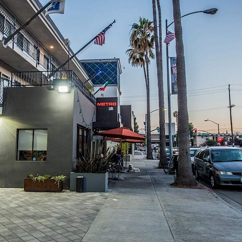 A street view at sunset with parked cars, buildings, a sidewalk café with umbrellas, palm trees, and flags. The sky is partially clear, ending the sentence.