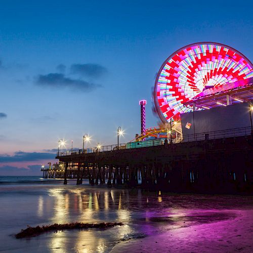 The image shows a brightly lit Ferris wheel and pier at dusk, with reflections on the water below and a cloudy blue sky in the background.