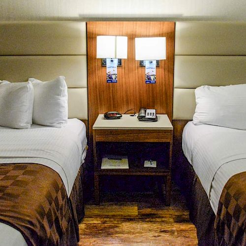 The image shows a hotel room with two neatly made beds, a nightstand in between with a lamp, a phone, and an alarm clock, and wooden flooring.