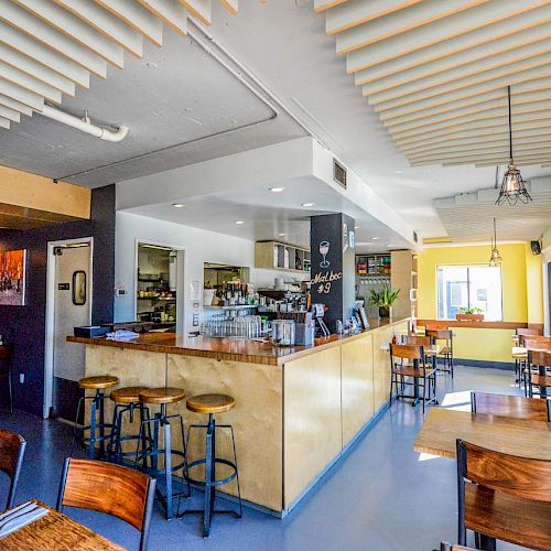 A modern, well-lit restaurant featuring wooden tables, chairs, and a bar area with stools, ready for patrons.