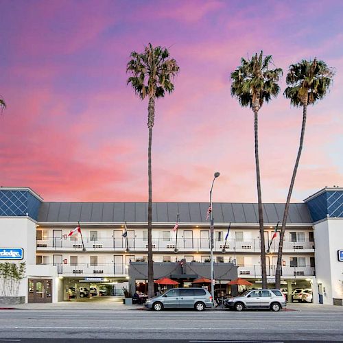 The image shows a three-story Travelodge hotel with palm trees in front, against a colorful sunset sky. There are cars parked in the front.