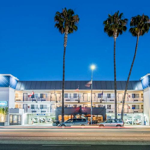 The image shows a well-lit hotel facade at dusk, with palm trees in front and some traffic passing on the road.