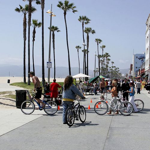 People are riding bicycles and walking along a busy boardwalk near the beach on a sunny day, with palm trees and shops lining the walkway.