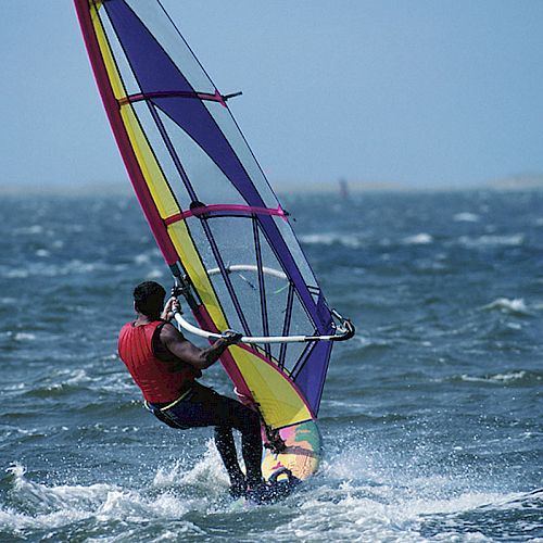 A person is windsurfing in choppy waters with a colorful sail, wearing a red vest. The background shows a cloudy sky and distant coastal landscape.