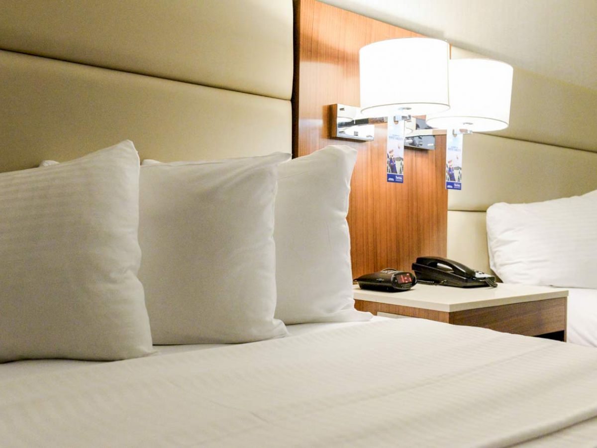 A neatly made bed in a hotel room, featuring three pillows, a bedside table with a telephone, and a wall-mounted lamp.