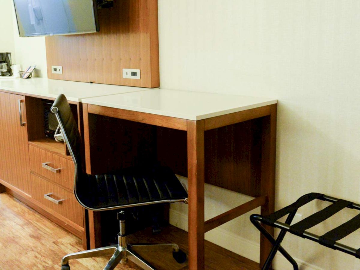 A minimalist hotel room setup featuring a TV, a wooden desk, an office chair, a coffee maker, storage drawers, and a luggage rack against a light wall.