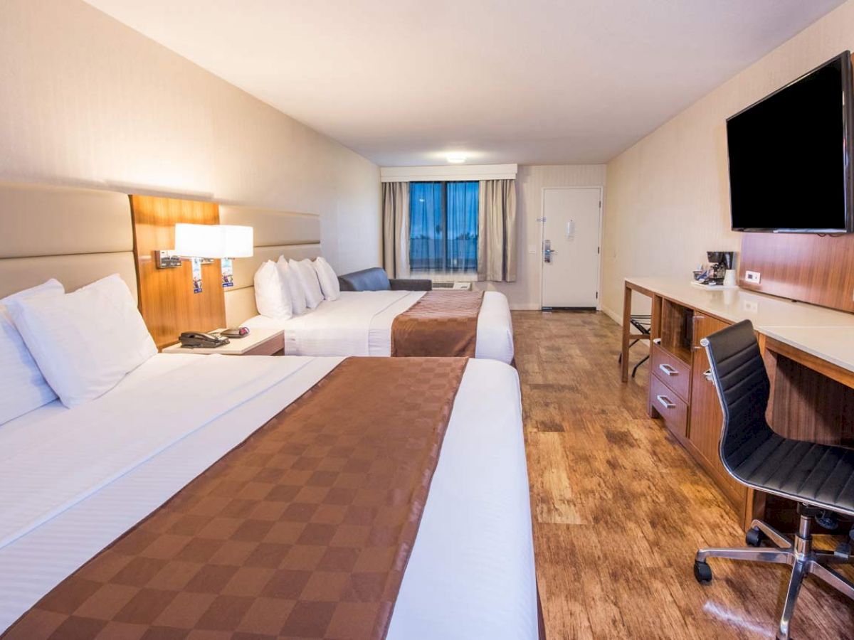 A hotel room with two beds, a desk, a chair, a TV, and a window in the background.