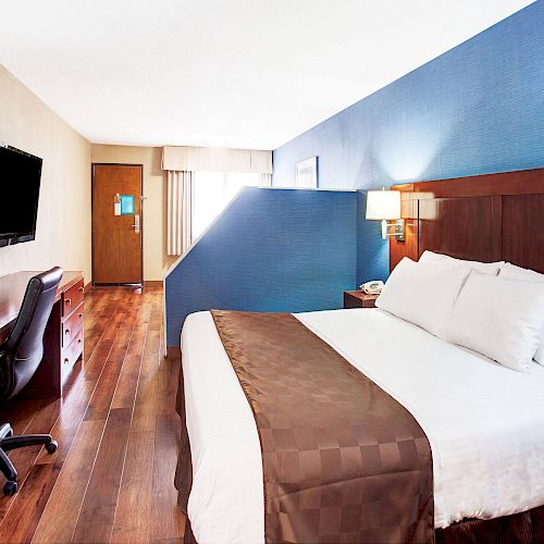 A hotel room with a large bed, a desk and chair, TV, lamps, and wooden flooring. The wall behind the bed is painted blue.