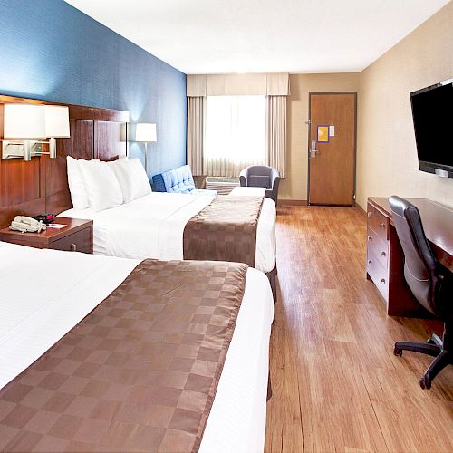 A hotel room with two double beds, desk, chair, flat-screen TV, bedside tables, lamps, wooden flooring, blue and beige walls, and a window.