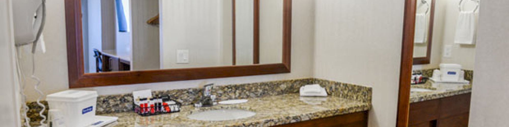 A bathroom vanity with a granite countertop, mirror, hairdryer, tissue box, toiletries, and towel rack on the wall, well-lit and clean.