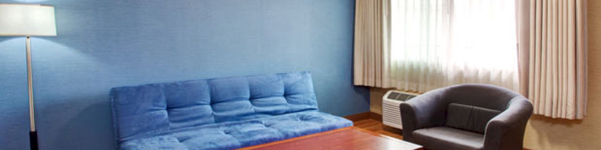 A room with a blue couch, a wooden coffee table, a dark armchair, a floor lamp, and a window with curtains.