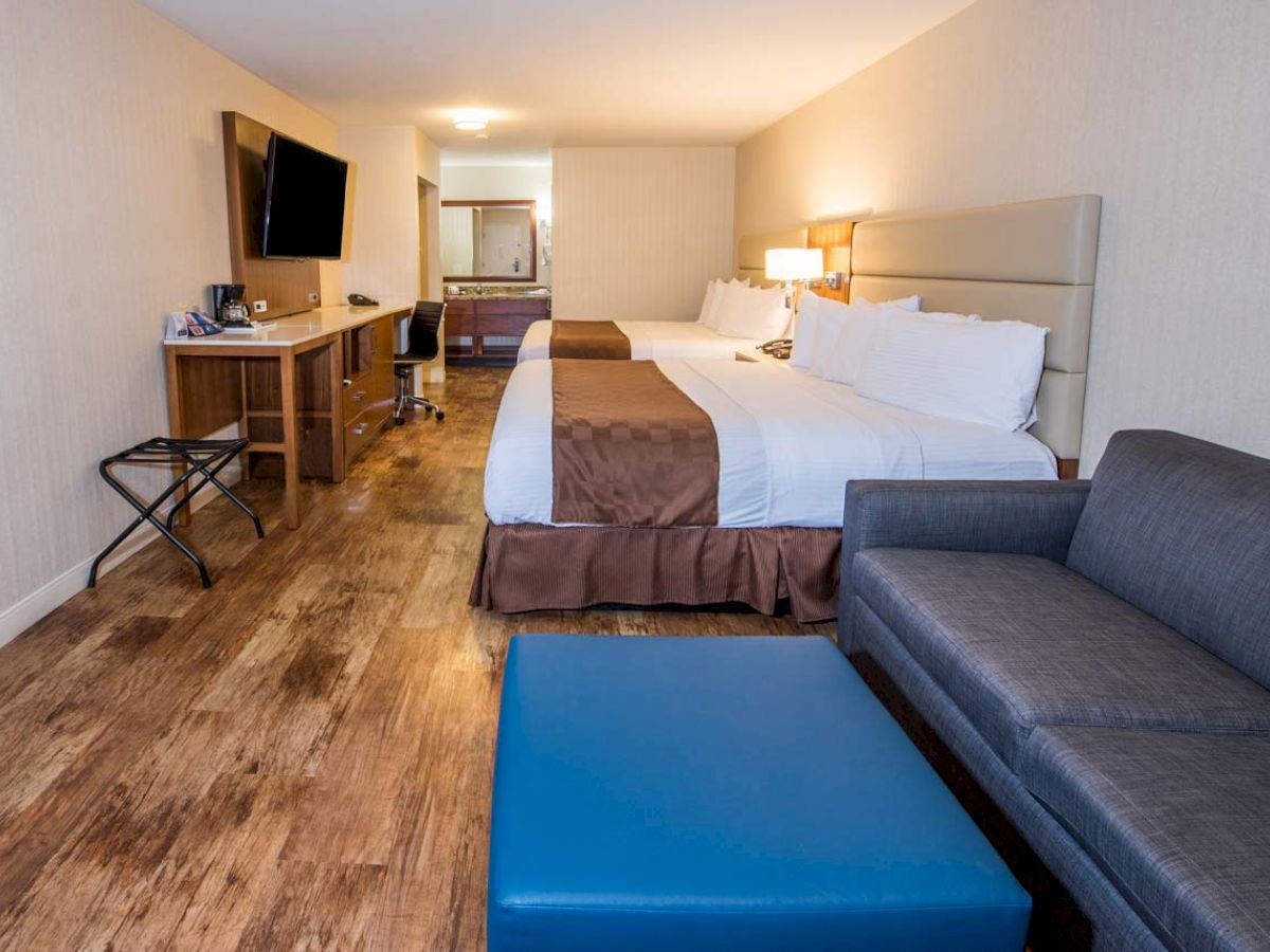 A hotel room with a large bed, sofa, blue ottoman, TV, desk, chair, and wooden floor, showing a cozy and modern design.