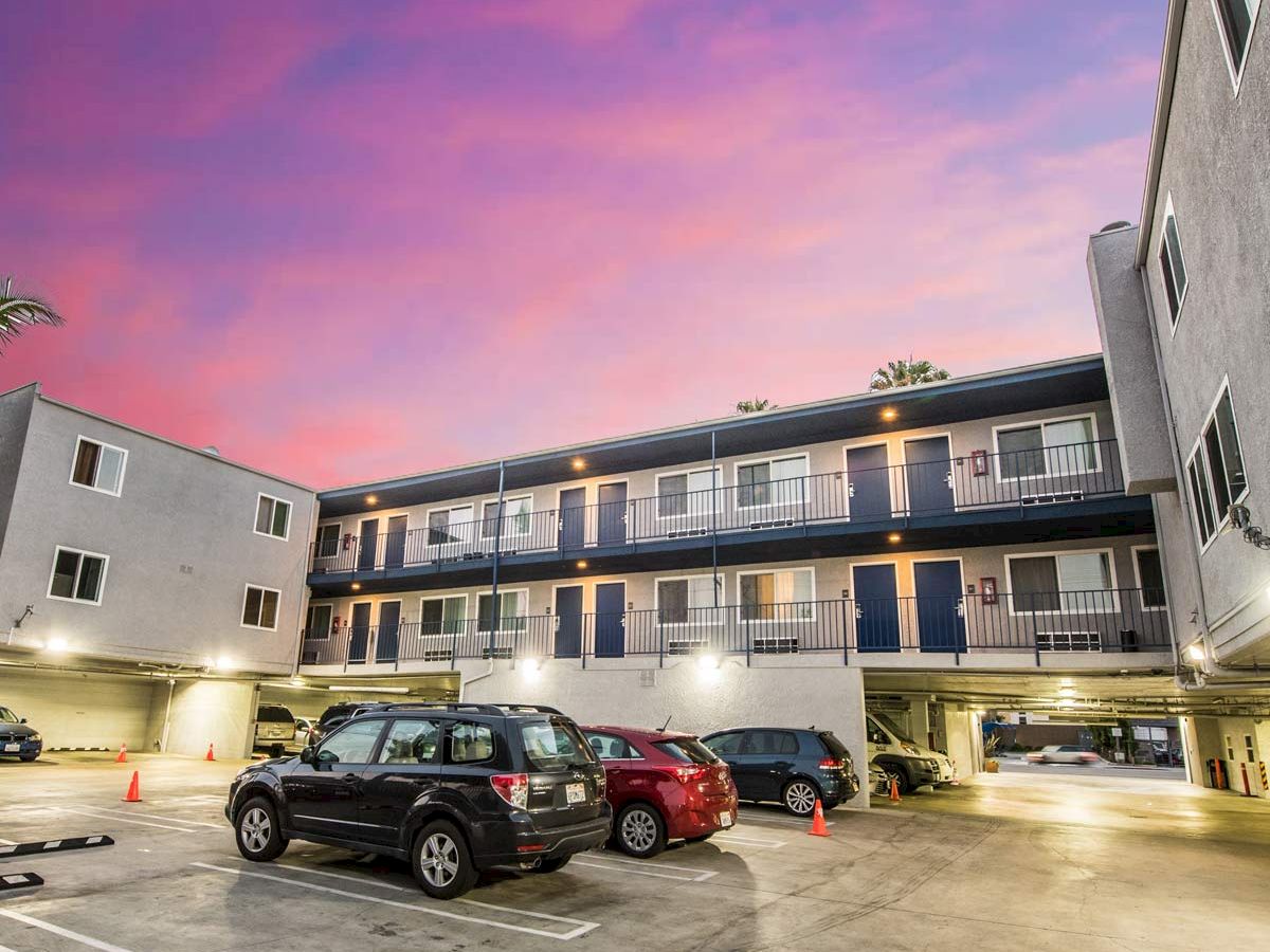 An apartment complex with a parking lot in front. Several cars are parked, and the sky is colorful with hues of pink and purple.