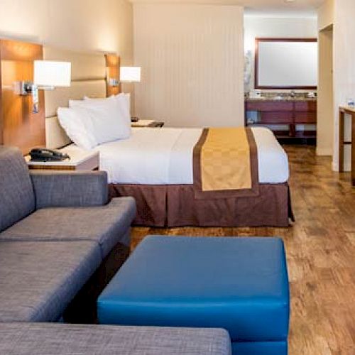 A hotel room with a double bed, sofa, blue ottoman, desk with chair, flat-screen TV, and modern lighting, featuring wood flooring and a bathroom area.