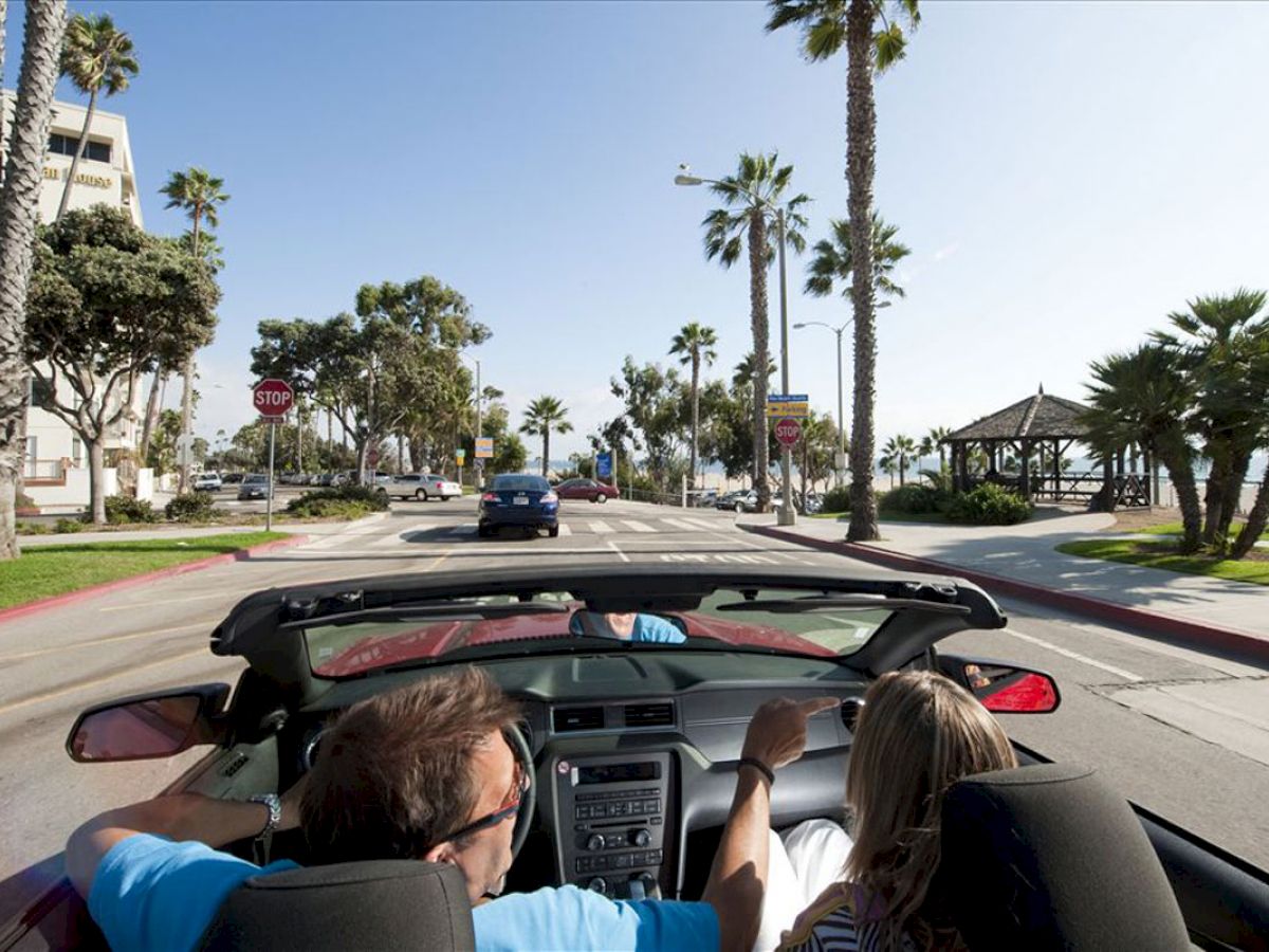 Two people are driving a convertible on a palm tree-lined street near the coast, enjoying a sunny day. The road is lined with palm trees.