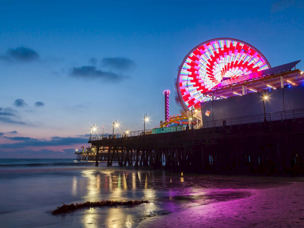 The image shows a night scene of a brightly lit Ferris wheel on a pier by the seaside, with its reflection shimmering on the water.