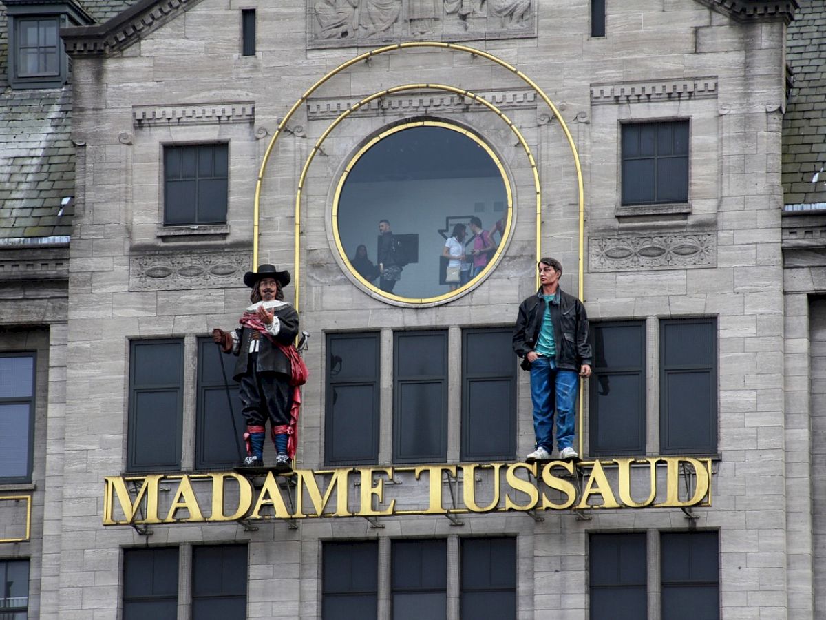 The image shows the facade of Madame Tussaud's museum with two wax figures on ledges outside and the museum’s name displayed prominently.